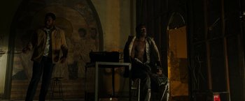 Movie still from “Candyman” (2021), directed by Nia DaCosta – A man sitting on a chair in a room; Wide shot, Low angle