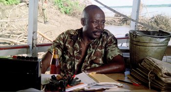 Movie still from “Cannibal Holocaust” (1980), directed by Ruggero Deodato – A man sitting at a table in front of a bottle of wine; Medium shot, Low angle