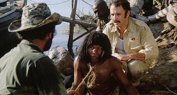 Movie still from “Cannibal Holocaust” (1980), directed by Ruggero Deodato – A group of people sitting around a body of water; Medium shot, Over the shoulder angle
