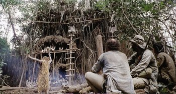 Movie still from “Cannibal Holocaust” (1980), directed by Ruggero Deodato – A man sitting on the ground in front of a fire pit; Wide shot, Low angle