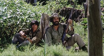 Movie still from “Cannibal Holocaust” (1980), directed by Ruggero Deodato – A man and a woman sitting in the grass holding guns; Medium shot, Low angle