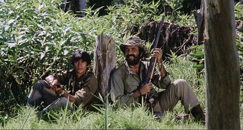 Movie still from “Cannibal Holocaust” (1980), directed by Ruggero Deodato – Two men sitting in the grass holding guns; Medium shot, Low angle