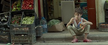 Movie still from “Capernaum” (2018), directed by Nadine Labaki – A young boy sitting on the sidewalk eating a sandwich; Wide shot, Over the shoulder angle