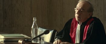 Movie still from “Capernaum” (2018), directed by Nadine Labaki – A judge sitting in front of a microphone and a glass bottle; Close Up shot, Over the shoulder angle