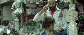 Movie still from “Capernaum” (2018), directed by Nadine Labaki – A man talking on a cell phone while a woman cuts a child's hair; Close Up shot, Over the shoulder angle