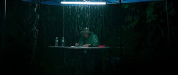 Movie still from “Cave Rescue” (2022), directed by Tom Waller – A man sitting under an awning in the rain writing; Wide shot, High angle