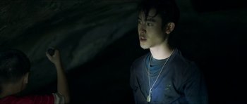 Movie still from “Cave Rescue” (2022), directed by Tom Waller – A young man wearing a necklace in a dark room; Close Up shot, Over the shoulder angle