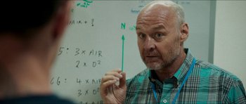 Movie still from “Cave Rescue” (2022), directed by Tom Waller – An older man holding a green stick in front of a whiteboard; Close Up shot, Over the shoulder angle