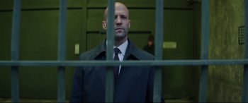 Movie still from “Fast & Furious Presents: Hobbs & Shaw” (2019), directed by David Leitch – A bald man in a black jacket behind a green fence; Medium shot, Low angle
