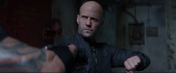 Movie still from “Fast & Furious Presents: Hobbs & Shaw” (2019), directed by David Leitch – A bald man in a black jacket is holding his hands out; Close Up shot, Over the shoulder angle
