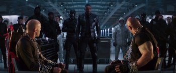 Movie still from “Fast & Furious Presents: Hobbs & Shaw” (2019), directed by David Leitch – A group of people in a room with a man in a black suit; Wide shot, Over the shoulder angle