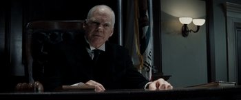 Movie still from “Changeling” (2008), directed by Clint Eastwood – An older man sitting in a chair in front of a flag; Medium shot, Low angle