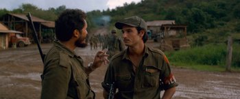 Movie still from “Che: Part One” (2008), directed by Steven Soderbergh – Two men smoking cigarettes while standing next to each other on a dirt road; Medium shot, Over the shoulder angle