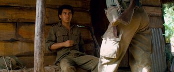 Movie still from “Che: Part One” (2008), directed by Steven Soderbergh – A man sitting in front of another man holding a rifle; Medium shot, Over the shoulder angle