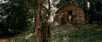 Movie still from “Che: Part One” (2008), directed by Steven Soderbergh – A man holding a rifle while standing next to a tree; Wide shot, Over the shoulder angle