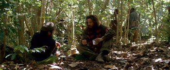 Movie still from “Che: Part One” (2008), directed by Steven Soderbergh – A man sitting on the ground in the woods; Medium shot, Over the shoulder angle
