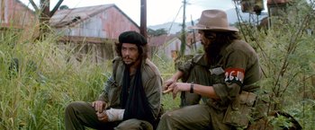 Movie still from “Che: Part One” (2008), directed by Steven Soderbergh – Two men sitting on the ground in the grass; Medium shot, Low angle