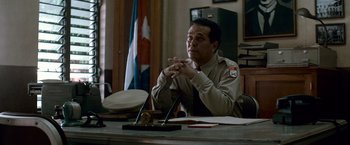 Movie still from “Che: Part One” (2008), directed by Steven Soderbergh – A man sitting at a table in front of a flag; Medium shot, Low angle