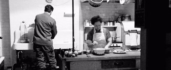 Movie still from “Che: Part One” (2008), directed by Steven Soderbergh – A woman in an apron preparing food in a kitchen; Medium shot, Over the shoulder angle
