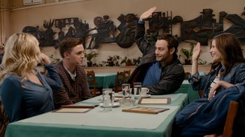 Movie still from “Chernobyl Diaries” (2012), directed by Bradley Parker – Two young men sitting at a table in front of a painting; Medium shot, Over the shoulder angle