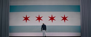 Movie still from “Chi-Raq” (2015), directed by Spike Lee – A man holding a baseball bat standing in front of a flag; Wide shot, Low angle