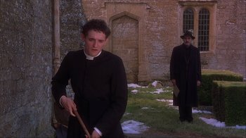 Movie still from “Chocolat” (2000), directed by Lasse Hallström – A man in a priest's outfit holding a baseball bat in front of an old building; Medium shot, Low angle