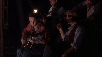 Movie still from “Chocolat” (2000), directed by Lasse Hallström – A group of men sitting on a bench playing a guitar; Medium shot, Low angle