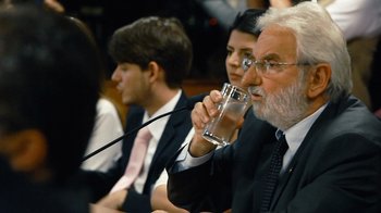 Movie still from “Citizenfour” (2014), directed by Laura Poitras – An older man drinking a glass of water while sitting in front of other people; Close Up shot, Over the shoulder angle