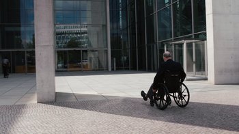 Movie still from “Citizenfour” (2014), directed by Laura Poitras – An older man sitting in a wheel chair outside of a building; Wide shot, High angle