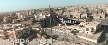 Movie still from “City of Ghosts” (2017), directed by Matthew Heineman – An aerial view of a city with a flag flying; Extreme Wide shot, High angle