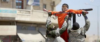 Movie still from “City of Ghosts” (2017), directed by Matthew Heineman – A man in orange is being held up by two soldiers in fatigues; Extreme Wide shot, Overhead angle