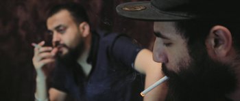 Movie still from “City of Ghosts” (2017), directed by Matthew Heineman – A man with a hat smoking a cigarette in front of another man; Close Up shot, Over the shoulder angle