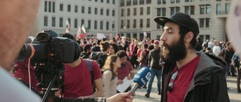Movie still from “City of Ghosts” (2017), directed by Matthew Heineman – A man is holding a microphone in front of a crowd of people; Medium shot, Over the shoulder angle