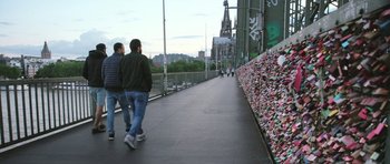 Movie still from “City of Ghosts” (2017), directed by Matthew Heineman – Two people walking down a sidewalk near a wall covered in locks; Extreme Wide shot, High angle
