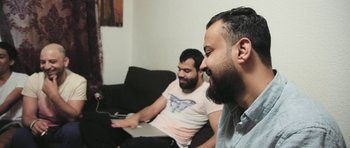 Movie still from “City of Ghosts” (2017), directed by Matthew Heineman – Two men sitting on a couch looking at something on a computer; Medium shot, Low angle