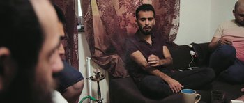 Movie still from “City of Ghosts” (2017), directed by Matthew Heineman – A man sitting on a couch smoking a hookah pipe; Medium shot, High angle