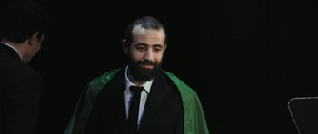 Movie still from “City of Ghosts” (2017), directed by Matthew Heineman – A man with a beard wearing a black suit and a green cape; Medium shot, Low angle
