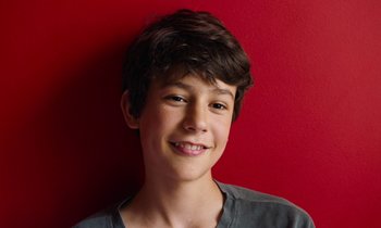 Movie still from “Close” (2022), directed by Lukas Dhont – A young boy smiling for the camera in front of a red wall; Close Up shot, Overhead angle