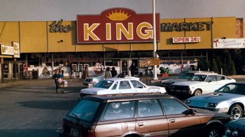 Movie still from “Cobra” (1986), directed by George P. Cosmatos – Cars are parked on the side of the road near a king market sign; Wide shot, Low angle