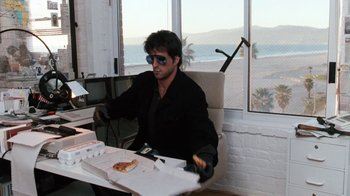 Movie still from “Cobra” (1986), directed by George P. Cosmatos – A man sitting in front of a box of pizza on top of a desk; Wide shot, Low angle