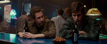 Movie still from “Colossal” (2016), directed by Nacho Vigalondo – Two men sitting at a table with a bowl of pretzels and a beer; Medium shot, Over the shoulder angle