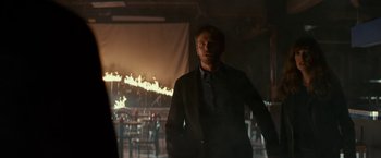 Movie still from “Colossal” (2016), directed by Nacho Vigalondo – A man standing in a room with a fire in the background; Medium shot, Over the shoulder angle