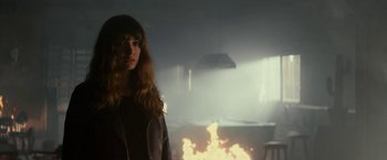 Movie still from “Colossal” (2016), directed by Nacho Vigalondo – A woman standing in front of a fire in a room; Medium shot, Over the shoulder angle