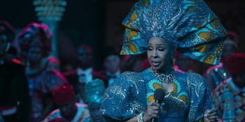 Movie still from “Coming 2 America” (2021), directed by Craig Brewer – A woman in a blue dress holding a microphone in front of a group of people; Close Up shot, Over the shoulder angle