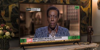 Movie still from “Coming 2 America” (2021), directed by Craig Brewer – A television screen showing a man in a suit and a necklace; Close Up shot, Over the shoulder angle