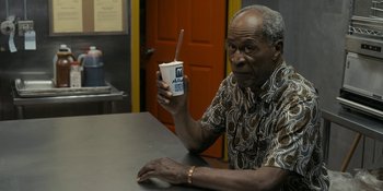 Movie still from “Coming 2 America” (2021), directed by Craig Brewer – An older man sitting at a table holding a drink; Medium shot, High angle