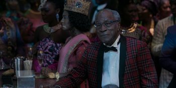 Movie still from “Coming 2 America” (2021), directed by Craig Brewer – An older man wearing a suit and bow tie in front of other people; Medium shot, Over the shoulder angle