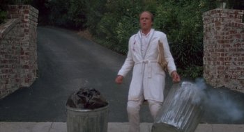 Movie still from “Commando” (1985), directed by Mark L. Lester – A man in a white bathrobe standing next to a trash can; Medium shot, Low angle