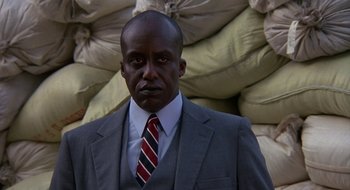 Movie still from “Commando” (1985), directed by Mark L. Lester – A man in a suit and tie standing in front of bags; Close Up shot, Low angle