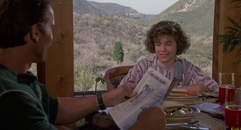 Movie still from “Commando” (1985), directed by Mark L. Lester – A person sitting on a chair reading a newspaper; Medium shot, Over the shoulder angle